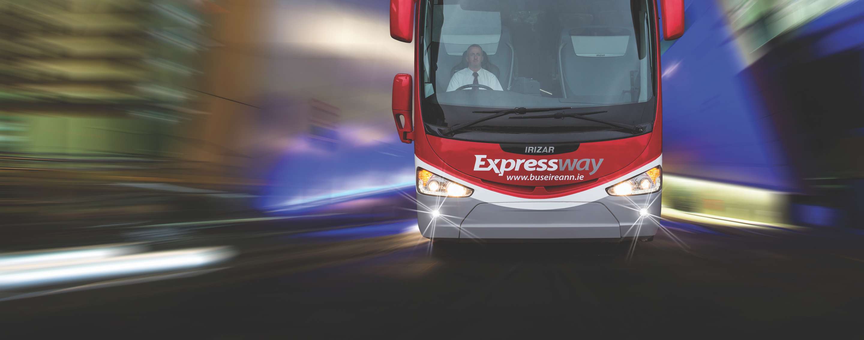Front view of a red-and-white Irizar “Expressway” coach of Bus Éireann, headlights on, driver in shirt and tie visible through the windshield, speeding past a blurred nighttime cityscape.